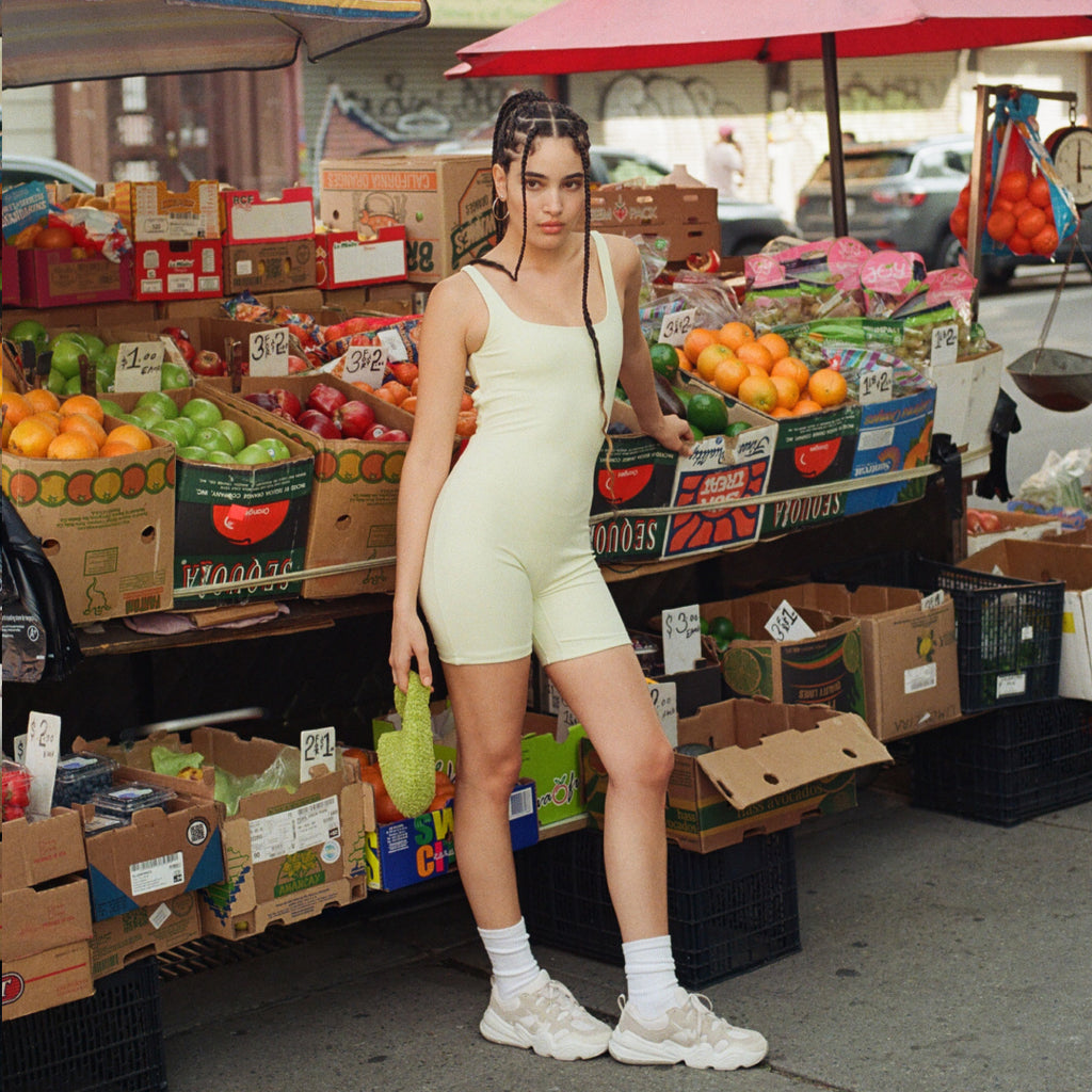 Model wearing the lime twist back catsuit from Le Ore in front of a fruit stand on the sidewalk in New York